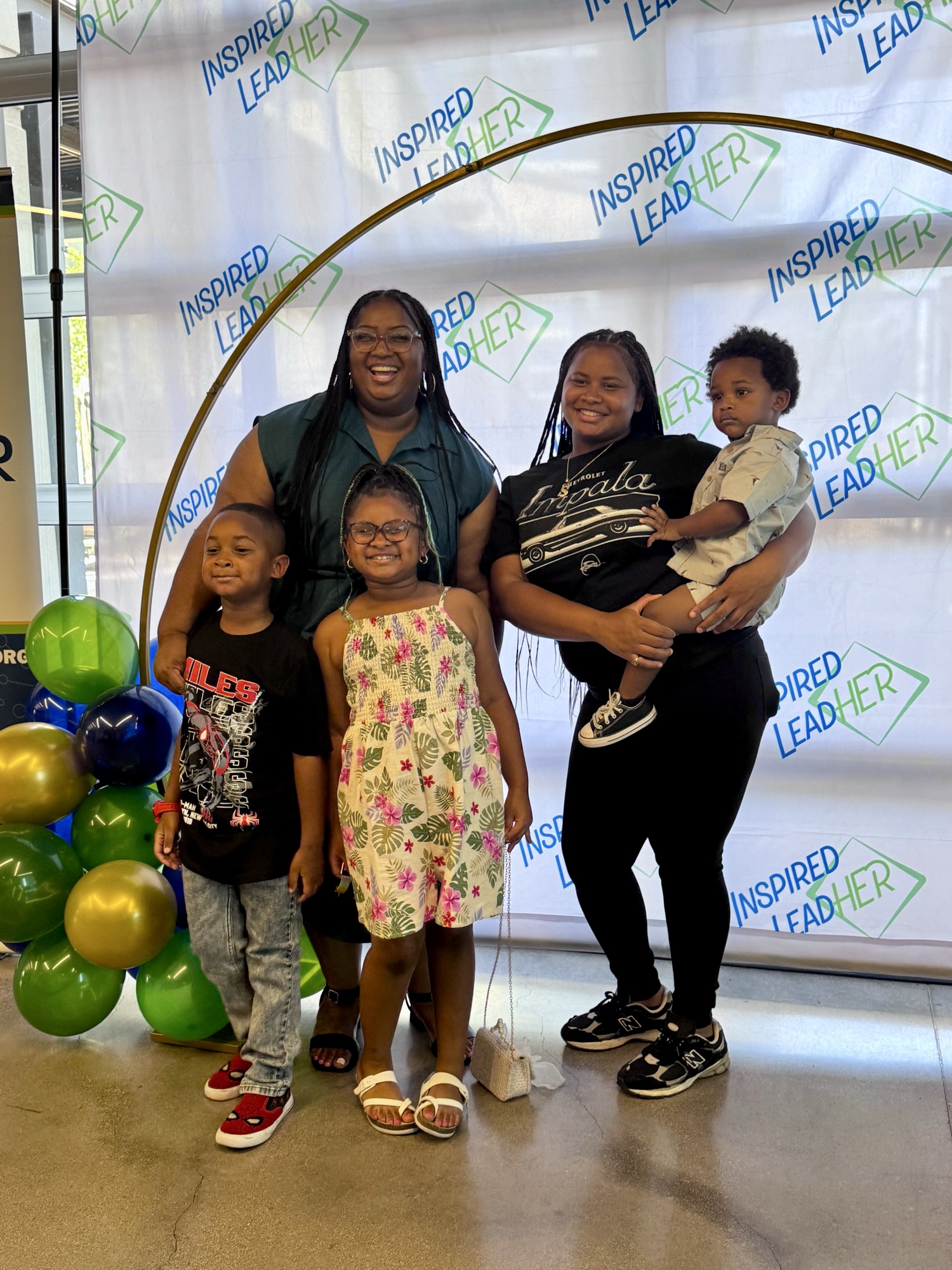 An Inspired LeadHER 2025 graduate posing with four family members, including children, under the gold balloon arch.